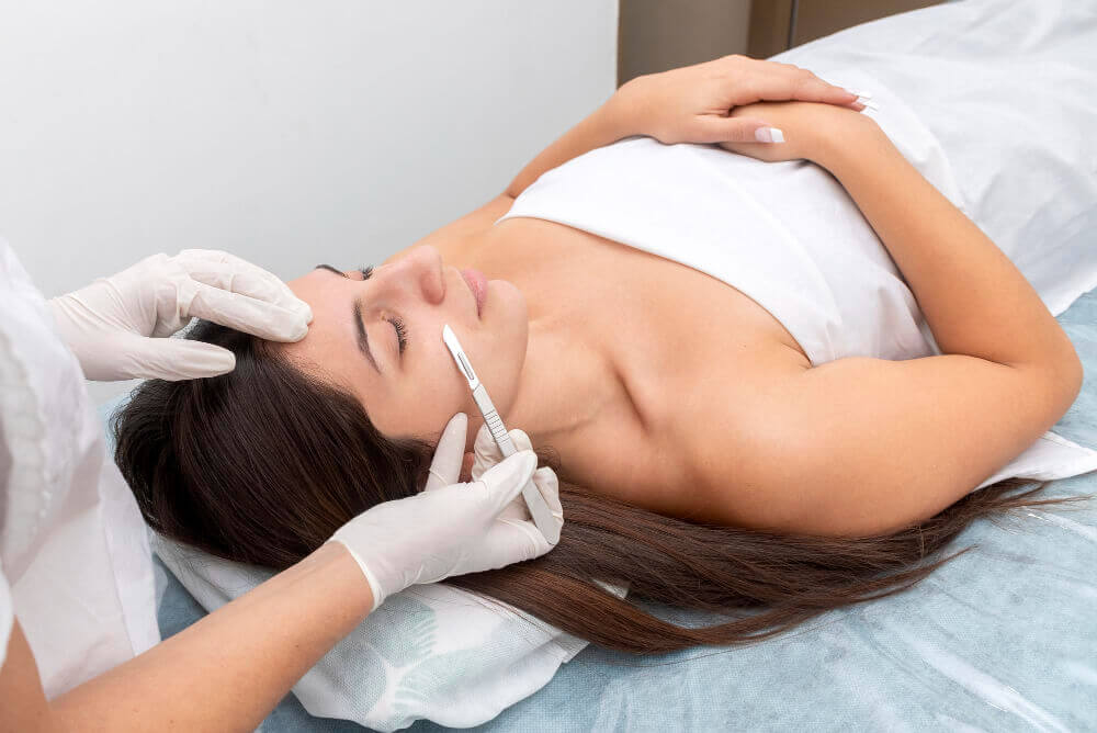 Professional performing dermaplaning facial with a sterile blade to remove dead skin and peach fuzz